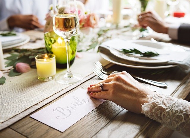 svatební hostina, nevěsta sedí u stolu, jde vidět ruka, která drží nápis bridge, dále je tam talíř, příbory, svíčka, sklenice a další věci